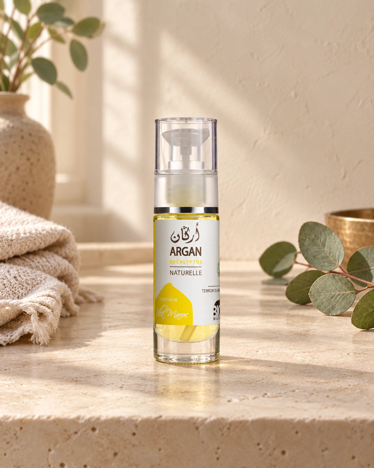 Huile d'Argan et d'Eucalyptus 30 ml sur une étagère en pierre claire, avec une branche d’eucalyptus, sous une douce lumière naturelle.