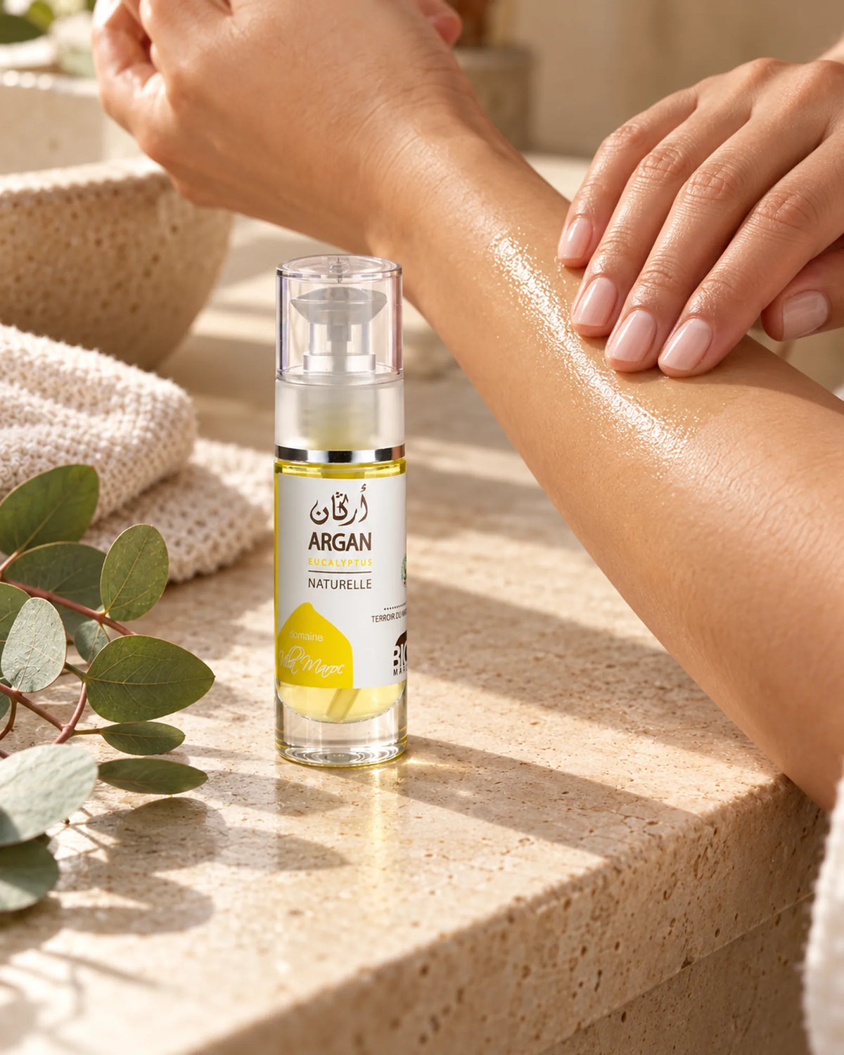 Huile d'Argan et d'Eucalyptus 30 ml sur une vasque en pierre, près de mains qui massent de l’huile sur un avant-bras.