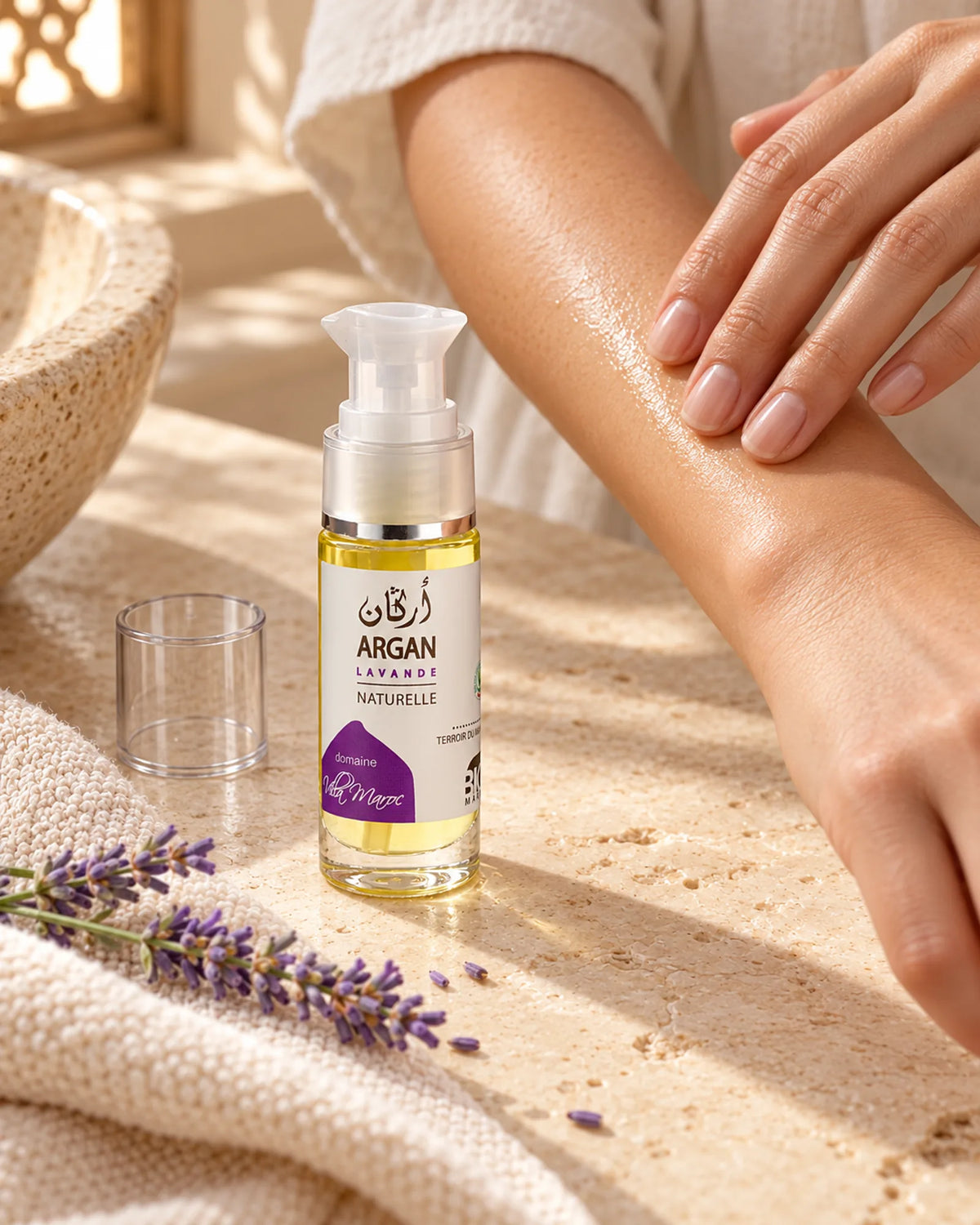 Huile d'Argan Lavande 30 ml sur une vasque en pierre, près de mains qui massent de l’huile sur un avant-bras.
